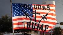 Large flag waves from a car, bearing the slogan "GOD, GUNS, TRUMP" with graphics of two rifles and a cross overlaid on the American flag stars and stripes.