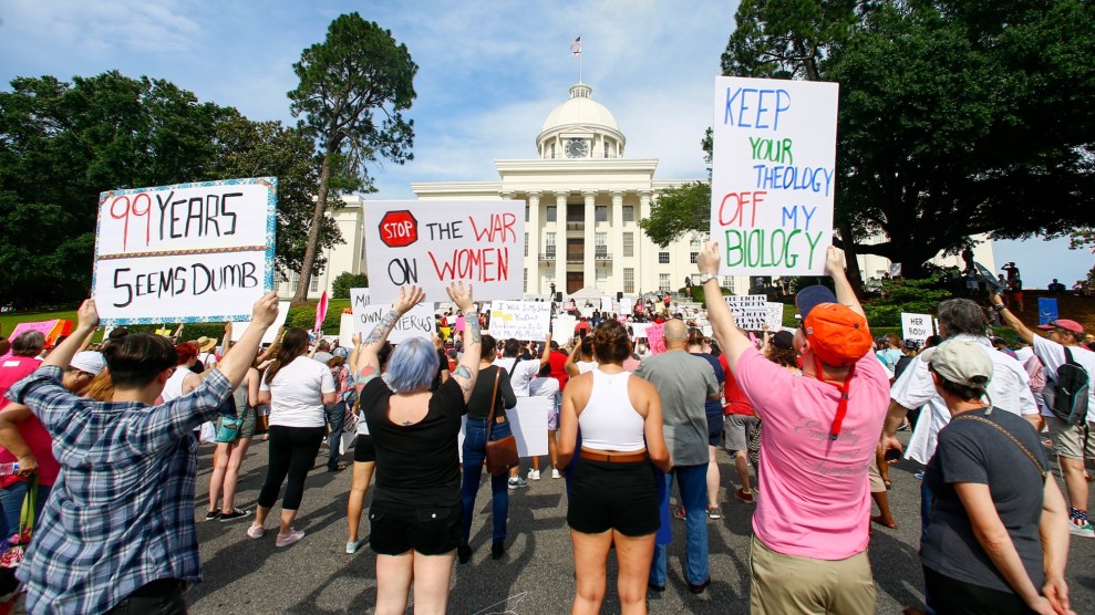 Seen from behind, a crowd of people face the Capitol. Signs held by protesters include, "99 years seems dumb," "Stop the war on women," and "Keep your theology off my biology."