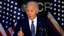 Joe Biden onstage in a navy suit, pointing with one of his fingers. Two American flags stand behind him.
