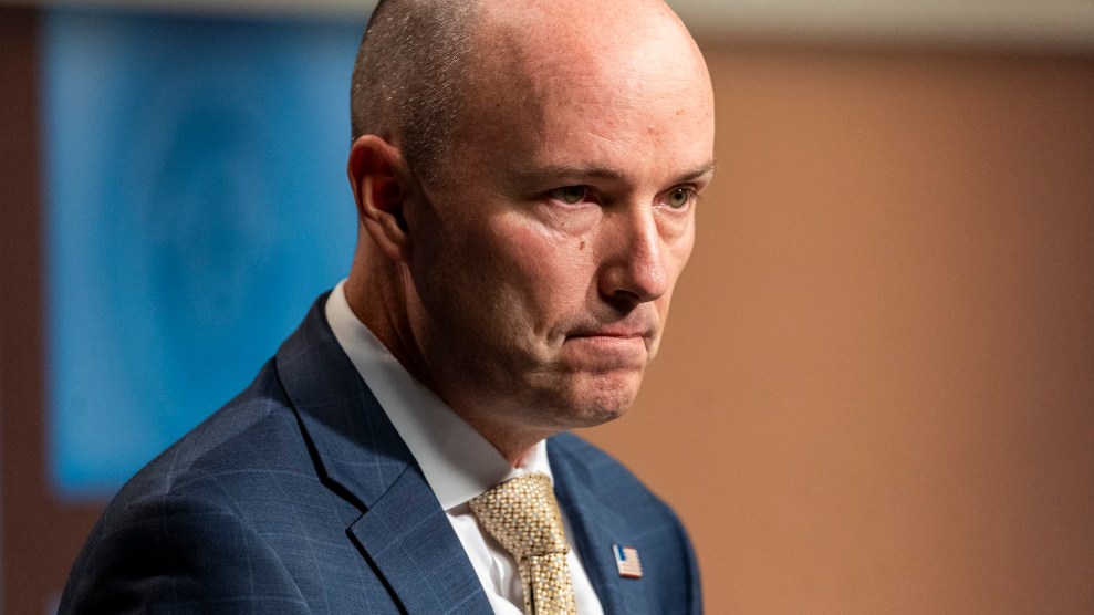 A mostly bald white man pursing his lips in a dark suit and yellow tie with an American flag lapel pin.